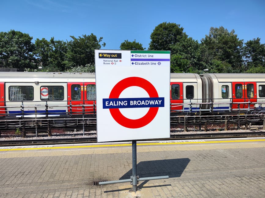 A clear view of a railway station platform with a prominent Ealing Broadway sign mounted on a freestanding metal post in the foreground. Behind the sign, a modern train with a silver body and red accents along the windows is parked on the tracks. The train's surface appears smooth with reflective glass windows, some of which reveal interior details. The platform surface is paved with textured light grey tiles, casting a shadow from the sign onto it. In the background, lush green treetops extend across the scene, contrasting with the urban elements. Above, a bright blue sky indicates fair weather. Nearby signage, partially visible, directs passengers to various lines and exits, typical of a public transit environment. The scene captures the tidy, organized appearance of an active railway station, which can relate to the broader context of transportation options for waste logistics, such as the potential use of private transport or independent collection methods for rubbish removal, aligned with services like those offered by House Clearance Ealing.