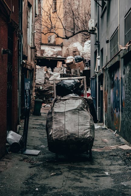 A narrow urban alleyway filled with a large pile of assorted rubbish and debris, including cardboard boxes, black trash bags, and loose waste materials stacked against a makeshift barrier at the end of the alley. In the foreground, a weathered metal trolley or wheeled bin with a worn, canvas-like cover is positioned on the asphalt surface, which shows signs of staining and dirt accumulation. The surrounding environment features graffiti-covered metal fencing on the right side and aged brick walls on the left, with some graffiti tags visible. Bare tree branches extend overhead, and the background reveals residential buildings with muted, faded facades. The scene is illuminated by natural light, casting subtle shadows, and overall suggests an area in need of rubbish removal or clearance, an activity often addressed by private waste management services like House Clearance Ealing performing on-site waste collection or disposal tasks in urban settings.