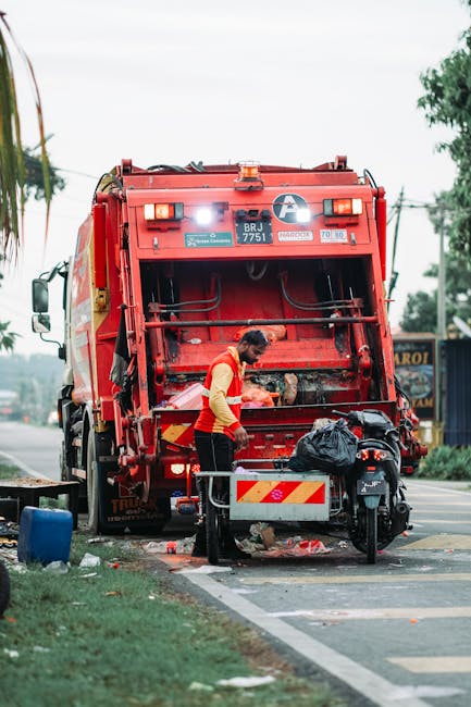 A worker dressed in a high-visibility orange vest and dark trousers is operating a large, red refuse collection vehicle on a paved street, engaged in waste collection activities. The back of the vehicle is open, revealing the internal mechanisms and storage area, with some debris and waste materials visible around the area. A black trash bag is placed on a small metal platform attached to the side of the truck, which also features reflective yellow and red warning markings on its rear. In the foreground, part of a motorcycle or scooter is parked beside the worker, with a small container or bin nearby. The surrounding environment includes a sidewalk with a grassy verge and some trees, utility poles with wires, and distant buildings, giving an impression of an urban or suburban setting. The sky is overcast, providing diffuse lighting that highlights the details of the waste removal scene, which is in alignment with private or independent rubbish collection services like those offered by House Clearance Ealing, with emphasis on efficient, professional waste handling in the context of residential or commercial clearance.