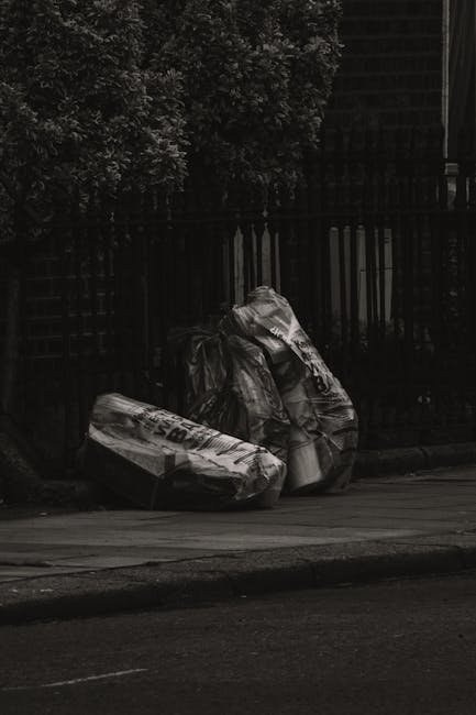 Two large black plastic bags filled with household waste and potentially bulky rubbish are placed on the pavement near the curb of a street, partially leaning against a black metal fence with vertical bars. The bags are crumpled and tightly packed, appearing to contain mixed refuse, and are situated close to a dense green hedge visible in the background. The scene suggests an informal waste collection method, typical of private rubbish removal services like those offered by House Clearance Ealing, especially when handling small loads or personal waste disposal outside regular council services. The environment appears to be a residential street, with the bags positioned on the edge of the footpath next to the roadway, under natural light, hinting at an outdoor setting suitable for independent rubbish collection or on-site clearance activities.