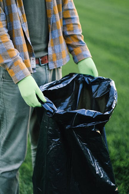 A person standing outdoors on a grassy area, wearing a casual plaid shirt with a green and gray checkered pattern, along with light green gloves, is holding open a large black rubbish bag made of thick plastic material. The bag is partially filled with waste and is positioned at waist height, ready for collection or disposal. The background shows a well-maintained lawn, suggesting a context of garden or property clearance. The scene emphasizes the process of individual waste management and private rubbish collection, aligning with services offered by companies like House Clearance Ealing that facilitate alternative waste handling solutions. The lighting appears natural, with soft daylight illuminating the scene, and the person's torso and hands are the central focus, with minimal environmental distractions, providing a clear visual connection to rubbish removal activities.