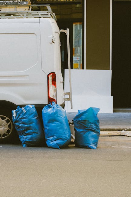 The image shows the rear section of a white commercial van parked on a street with three blue rubbish bags positioned on the pavement beside it, likely prepared for collection. The bags appear to be made of durable plastic with a slightly crinkled texture, and they are filled, causing them to bulge and stand upright. Part of the van's rear door and taillight are visible on the left side of the image, with the door slightly ajar, suggesting active loading or unloading. The background includes a storefront with large glass windows and doors, with white external paneling that partially covers the entrance. The scene is set during daylight, with natural lighting highlighting the clean, orderly appearance of the rubbish bags and vehicle. This image reflects a typical scene of private waste handling, potentially conducted by a rubbish removal service such as House Clearance Ealing, supporting efficient and alternative waste collection methods to local authority services.
