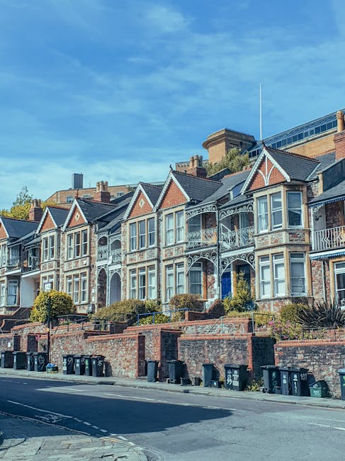 A row of Victorian-style terraced houses with brick facades, detailed white-painted ironwork balcony railings, and gabled roofs with slate tiles. The front gardens are elevated, separated from the street by low brick walls, with minimal landscaping including small shrubs and trees. The street scene is illuminated by natural daylight, showing a clear blue sky with some wispy clouds. At the bottom of the image, there are several black refuse bins lined up along the pavement, which are likely used for waste collection. The setting appears to be in a residential area where private waste disposal services, such as those provided by House Clearance Ealing, may be employed to manage rubbish removal or house clearance tasks, rather than relying solely on local authority collections. The overall scene emphasizes the importance of responsible waste handling in maintaining the appearance and cleanliness of the neighborhood.